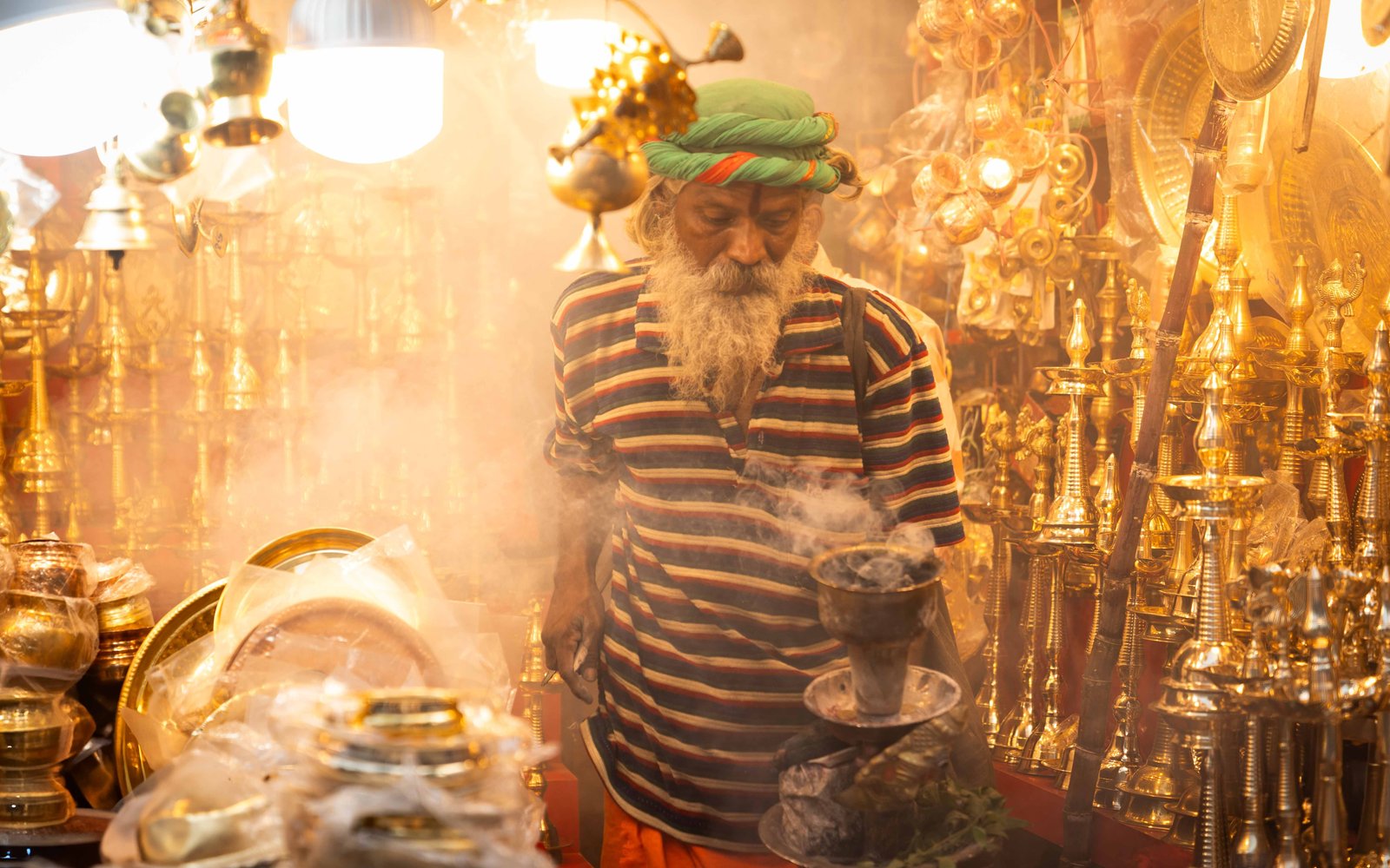 A man with a long white beard and navy and white striped top in a market setting amongst golden smoking pipes.