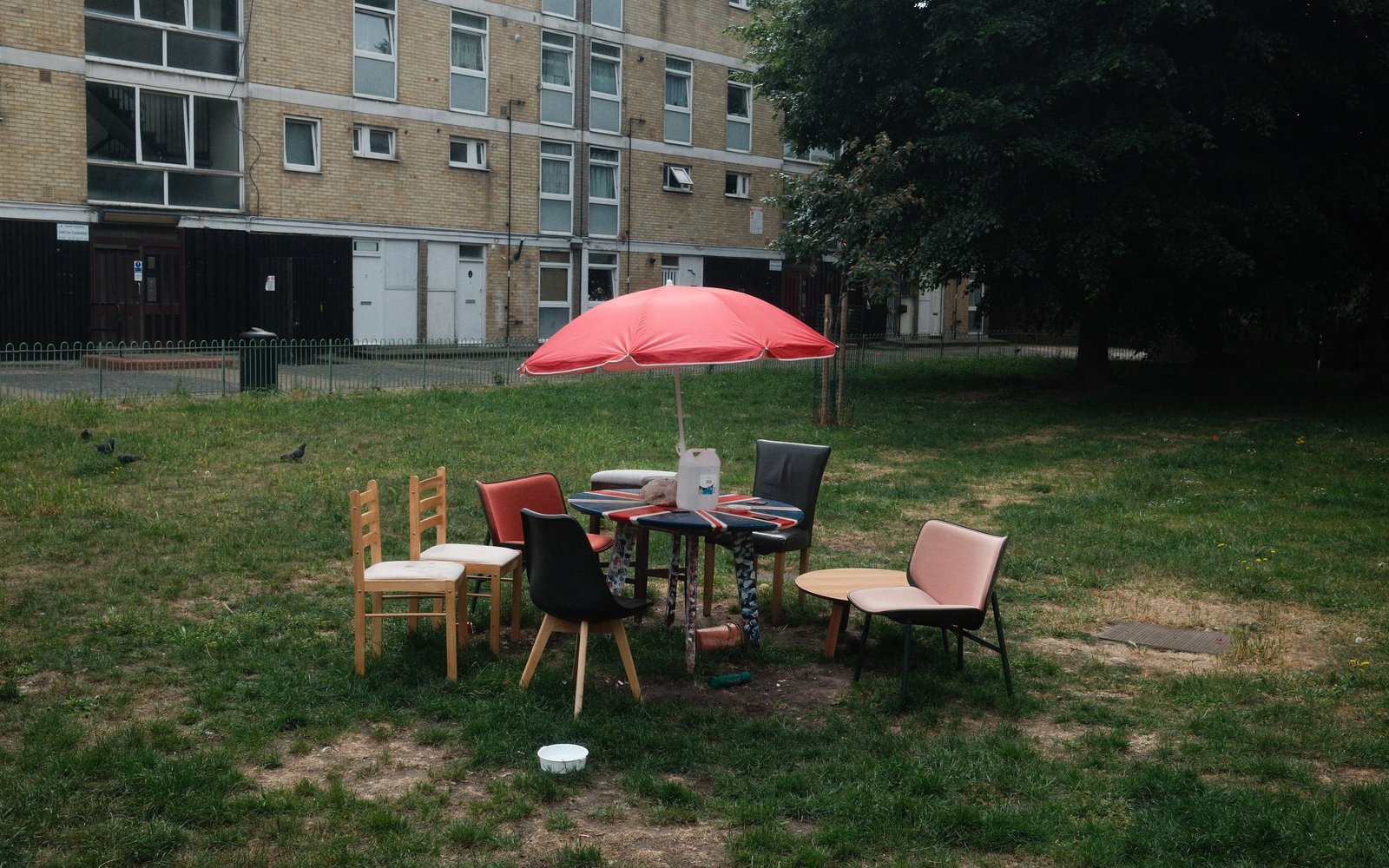 A table with umbrella and mismatched chairs on a lawn in front of an apartment block