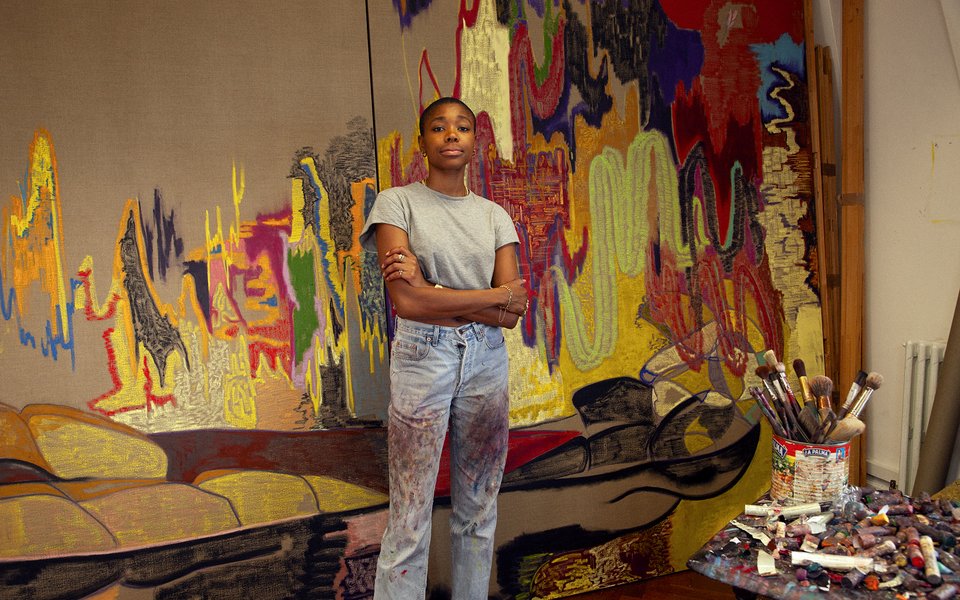 Photograph of a woman stood with hands on pocket looking at viewer with large abstract artwork behind her.