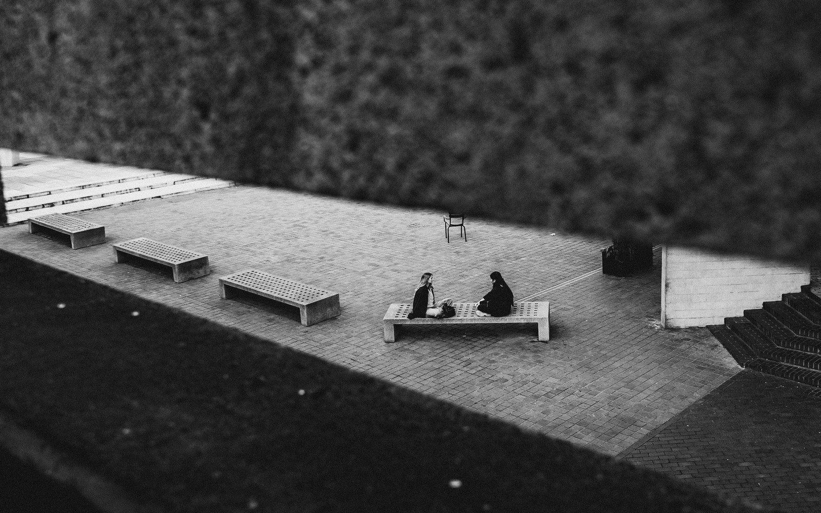 Two people sat on a bench with other benches in a row alongside, viewed through a gap in a balastrade outside