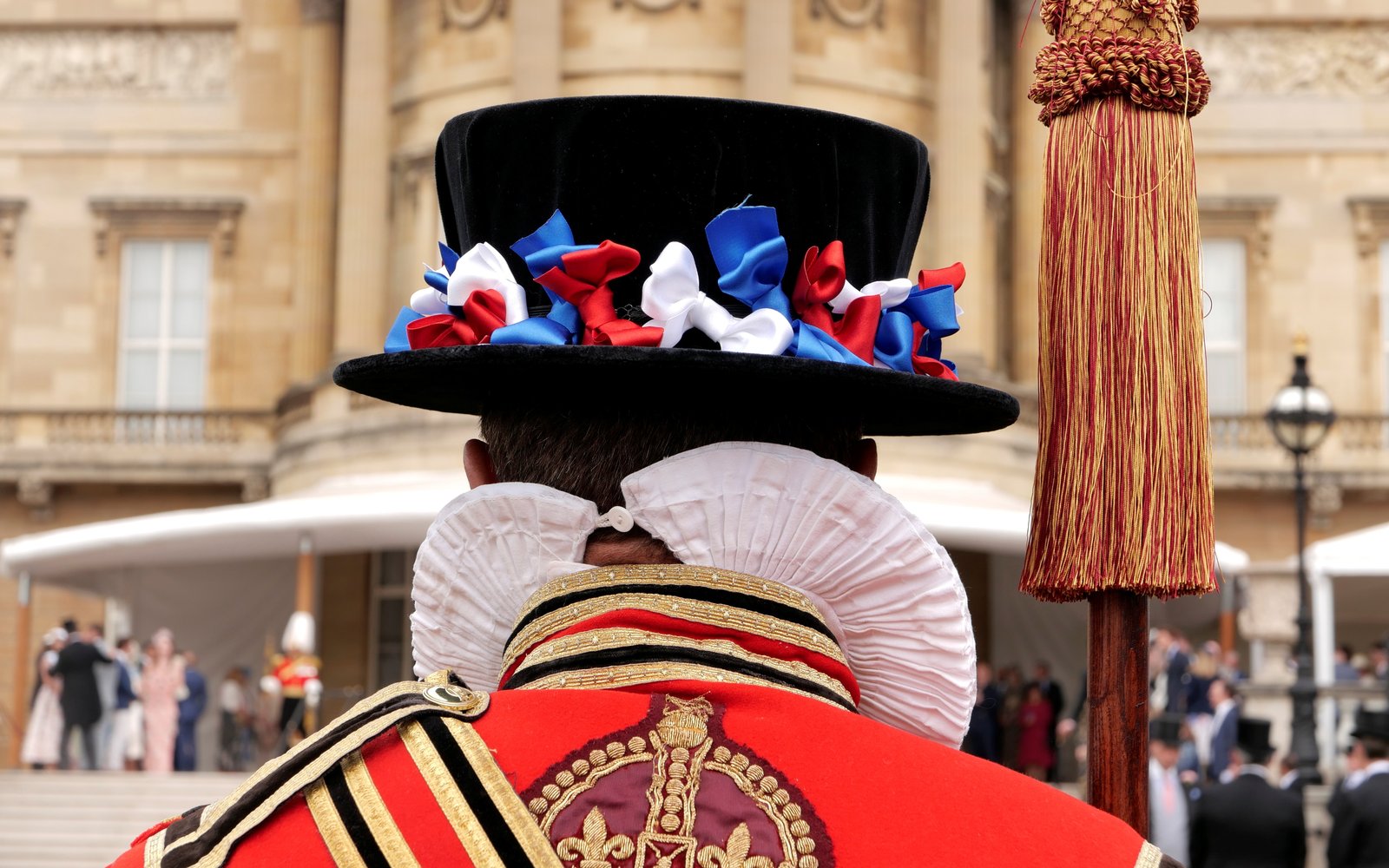 A beefeater pictured from behind wearing a black brimmed had with white red and blue ribbons, a white ruff and a red and gold traditional uniform.