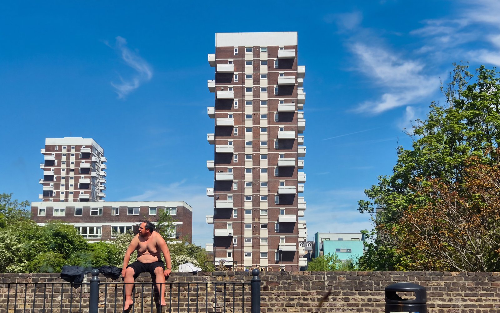 A man sat topless on a wall with a tower block behind against a bright blue sky.