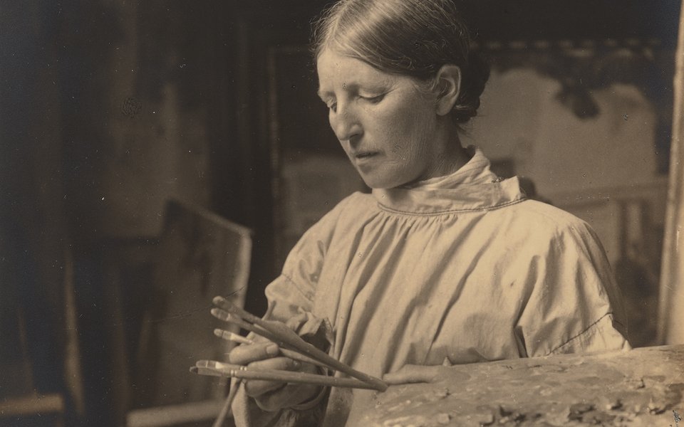Sepia photograph of a woman with hair tied back holding paintbrushes and a palette