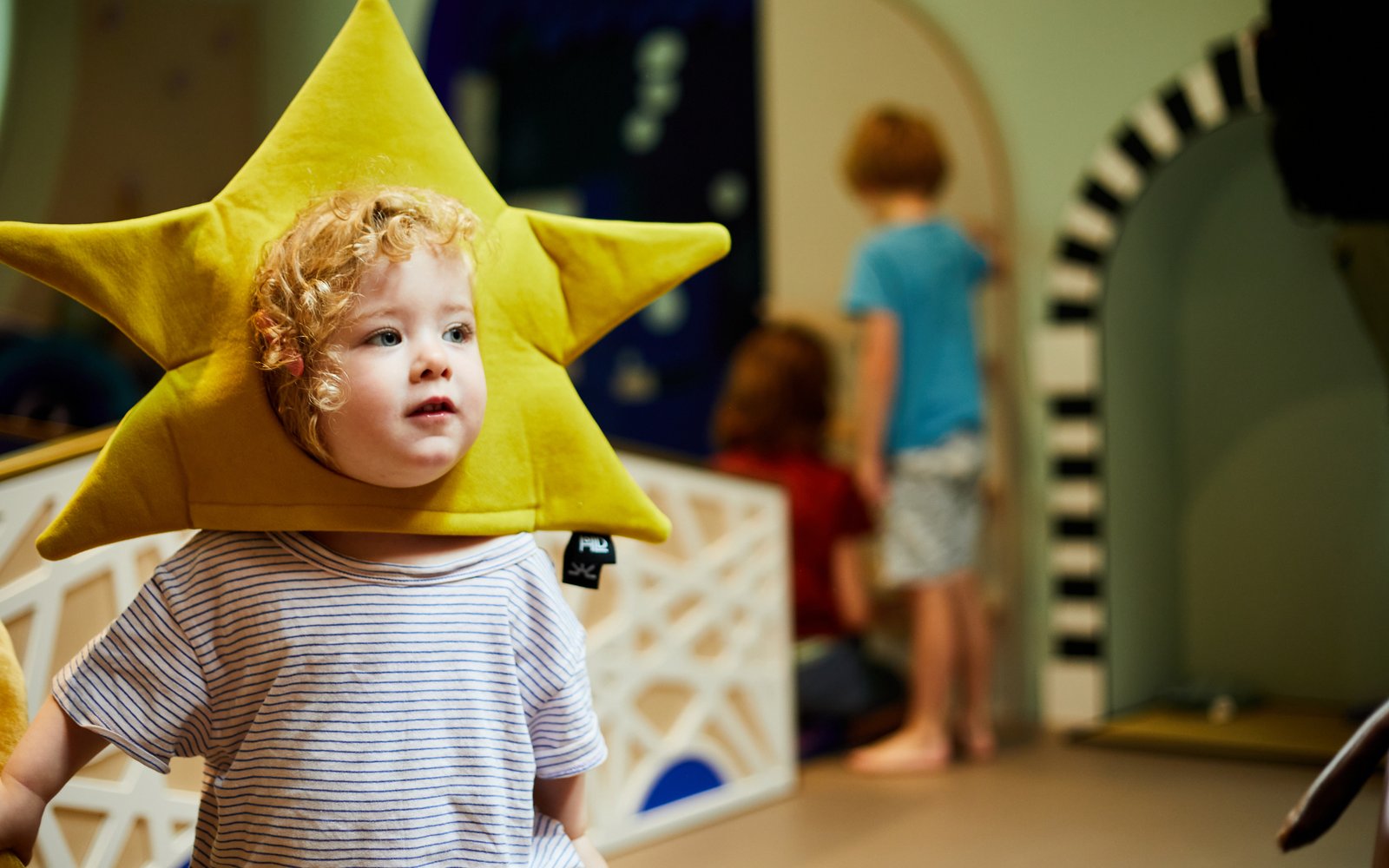 A young child with a colorful star-shaped hat, joyfully posing for a photo inside the ArtPlay Pavilion.