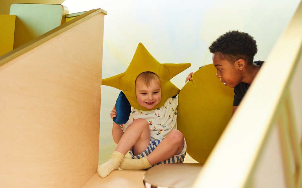 Two children at the top of a slide. One on left wears a star hat.