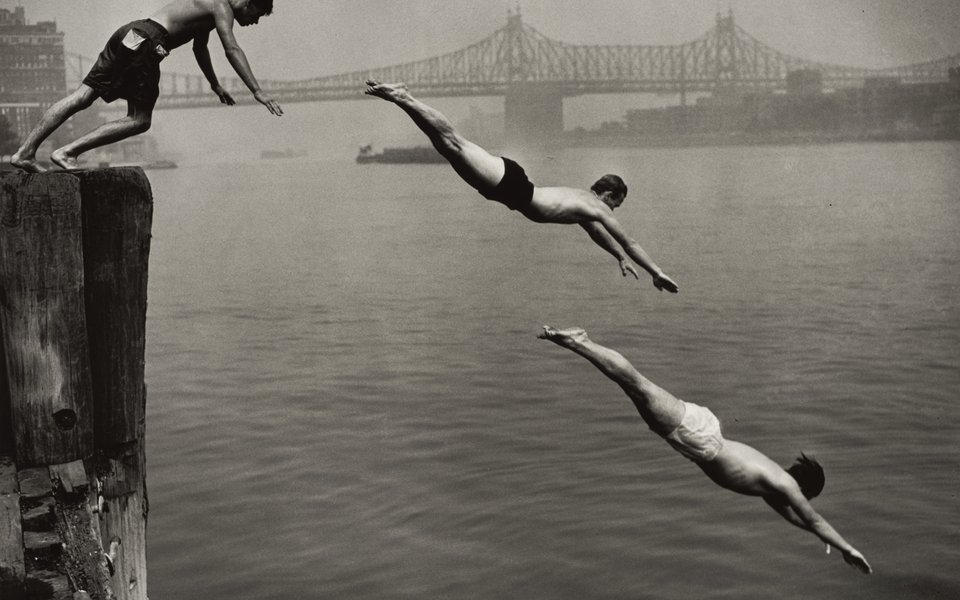 Three young boys diving into a river with a suspension bridge visible in the distance.