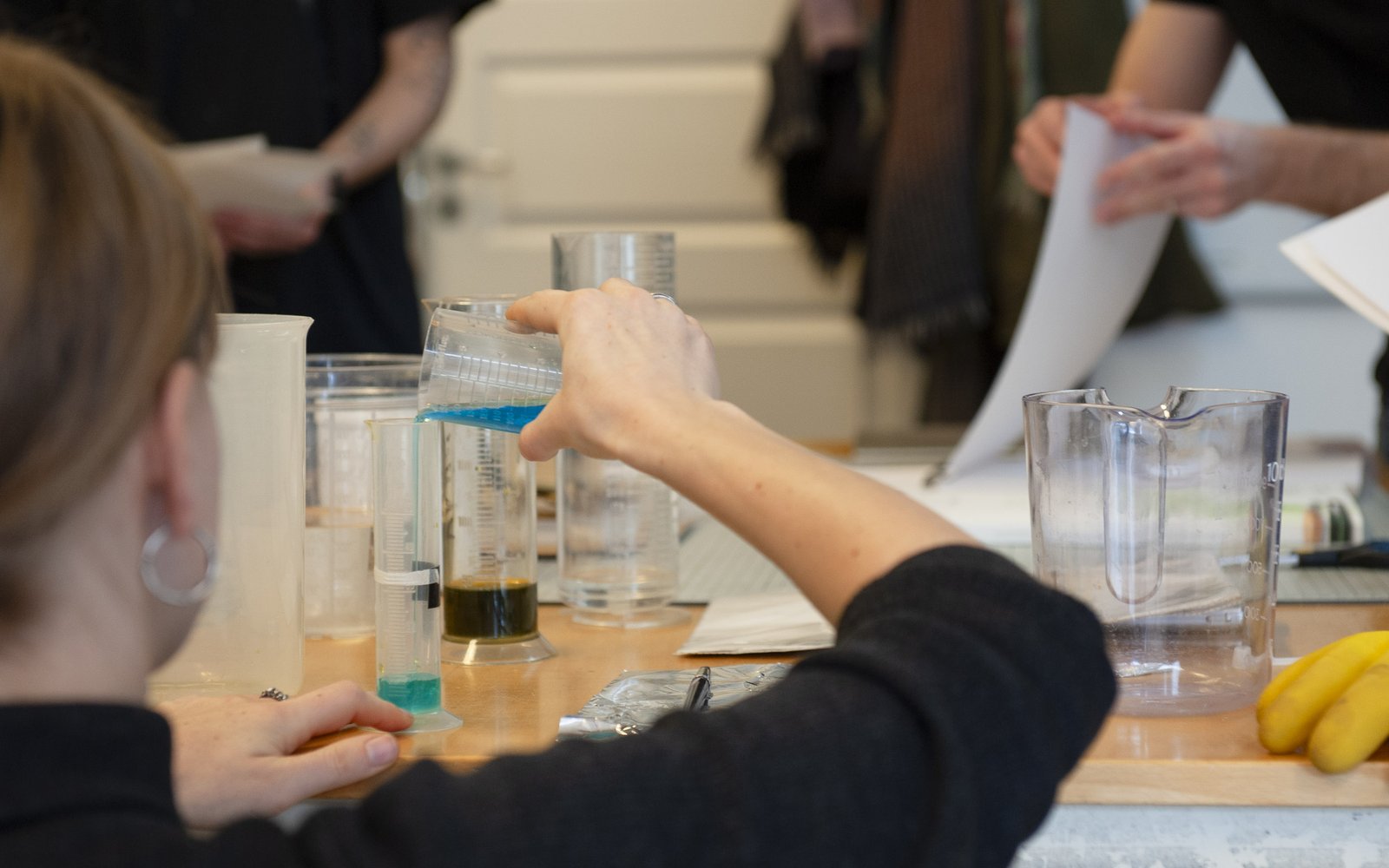 A woman at a table mixing chemicals in tall vessels while others look on