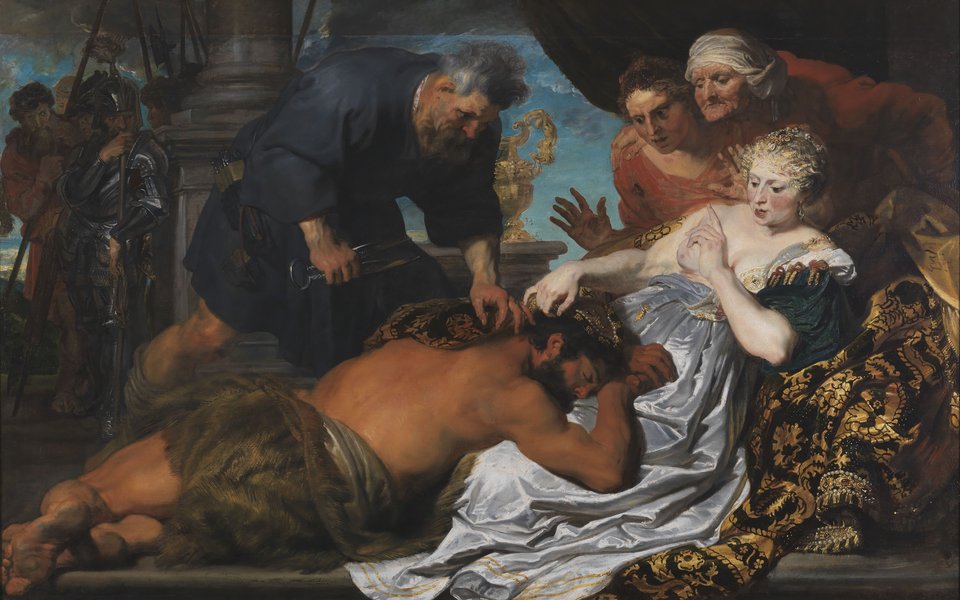 A man in a loinclothe lies in the lap of a woman in a blue silk dress with her breasts exposed. An older man holds a large scissors and is poised above the sleeping man about to cut his hair.