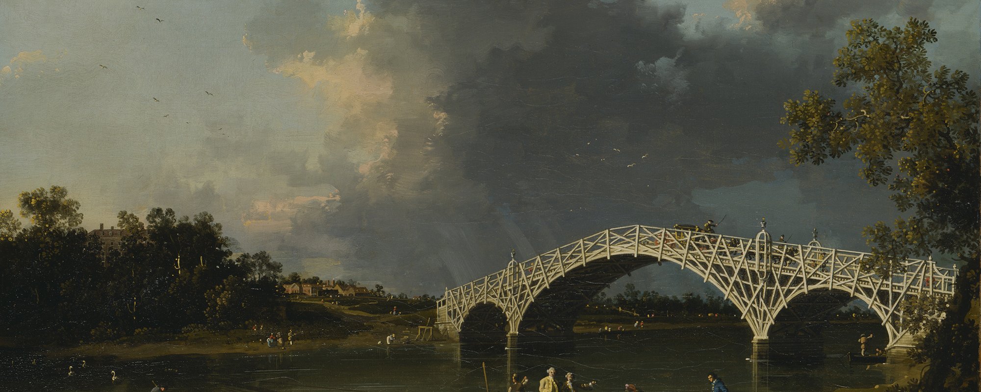 A white wooden arched bridge spans a calm river with people boating and walking nearby under a cloudy sky.