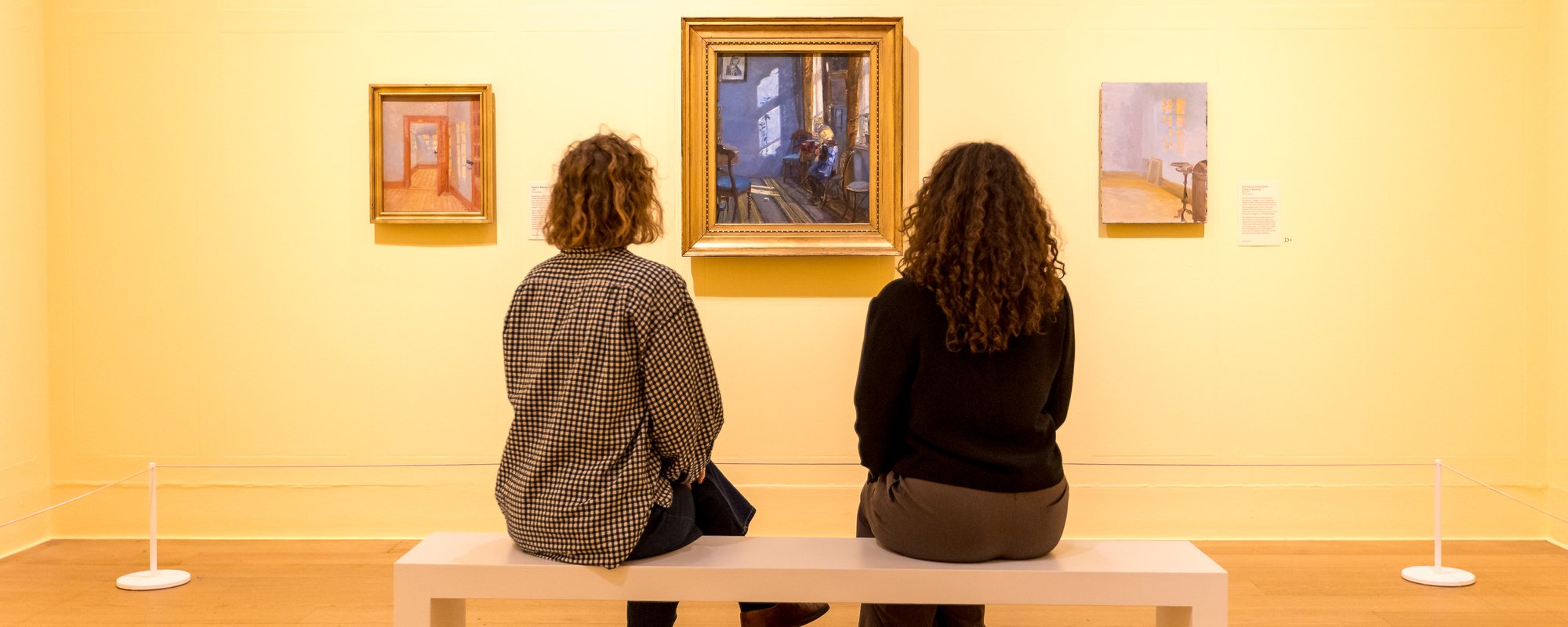 Two women sit on a white bench in an exhibition space looking at three paintings on a yellow wall.