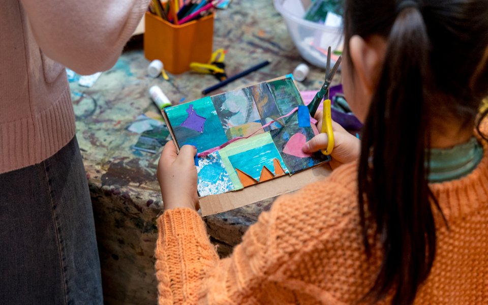 A child stands at a craft table with a scissors holding a card that she has made.