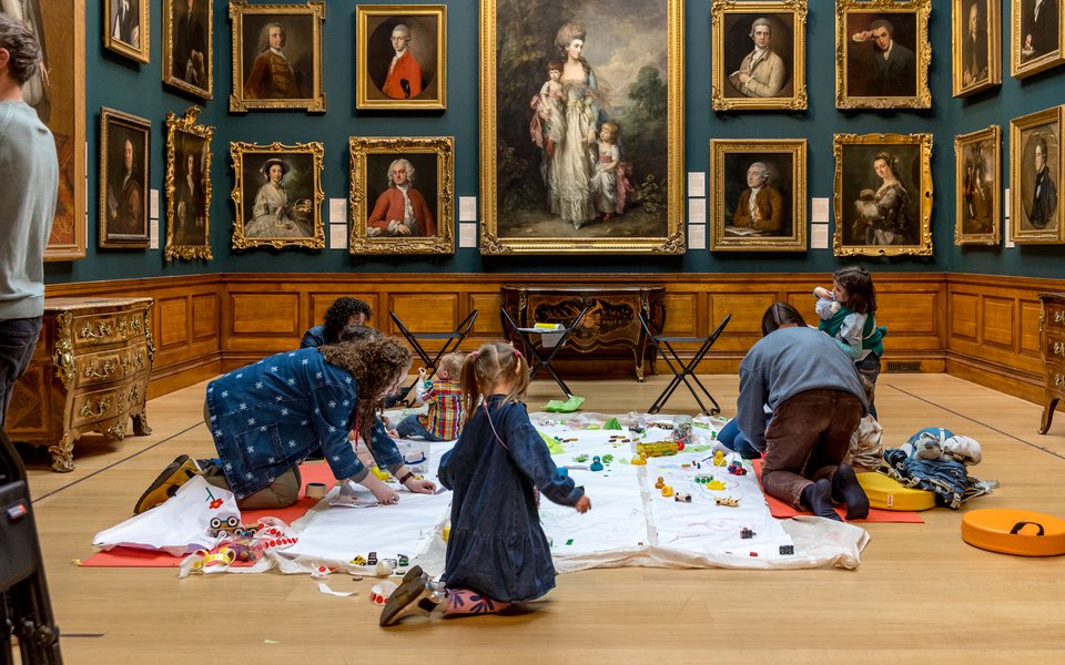 Family groups with small children play with toys on a mat on the floor of a gallery space with blue walls hung with gold framed historic portraits.