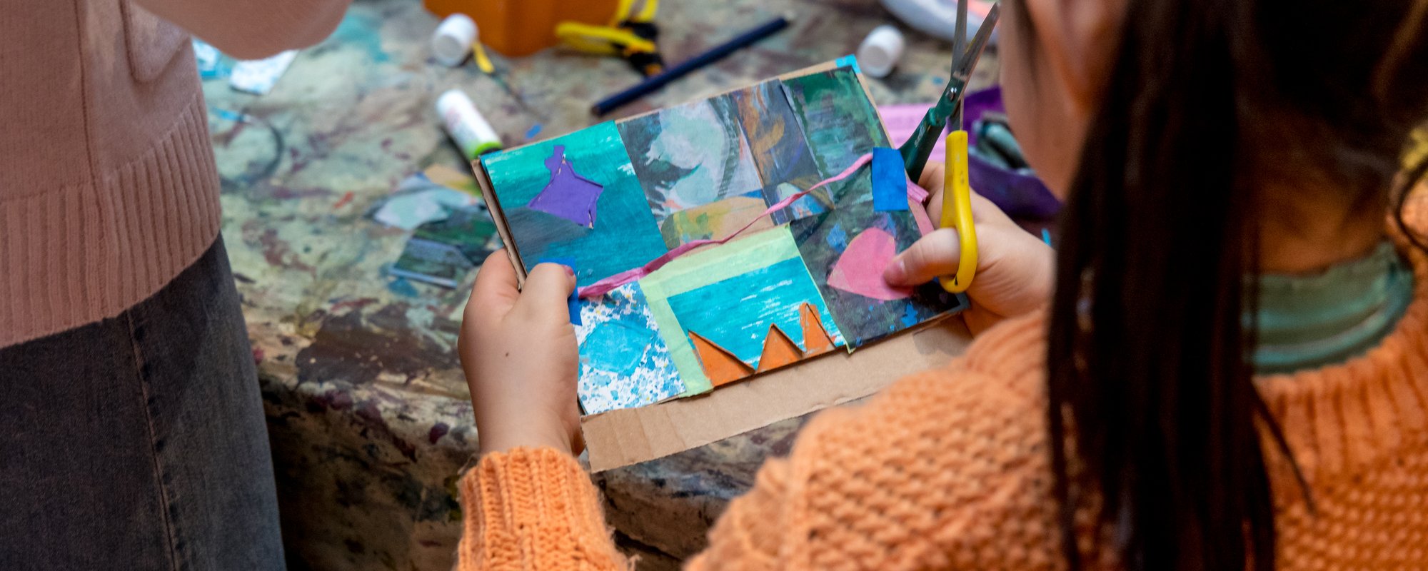 A child stands at a craft table with a scissors holding a card that she has made.