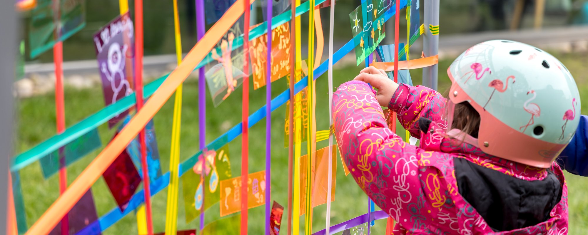 A young child wearing a pink coat and a blue helmet ats some ribbon to a lattice artwork of many different coloured ribbons and designs.
