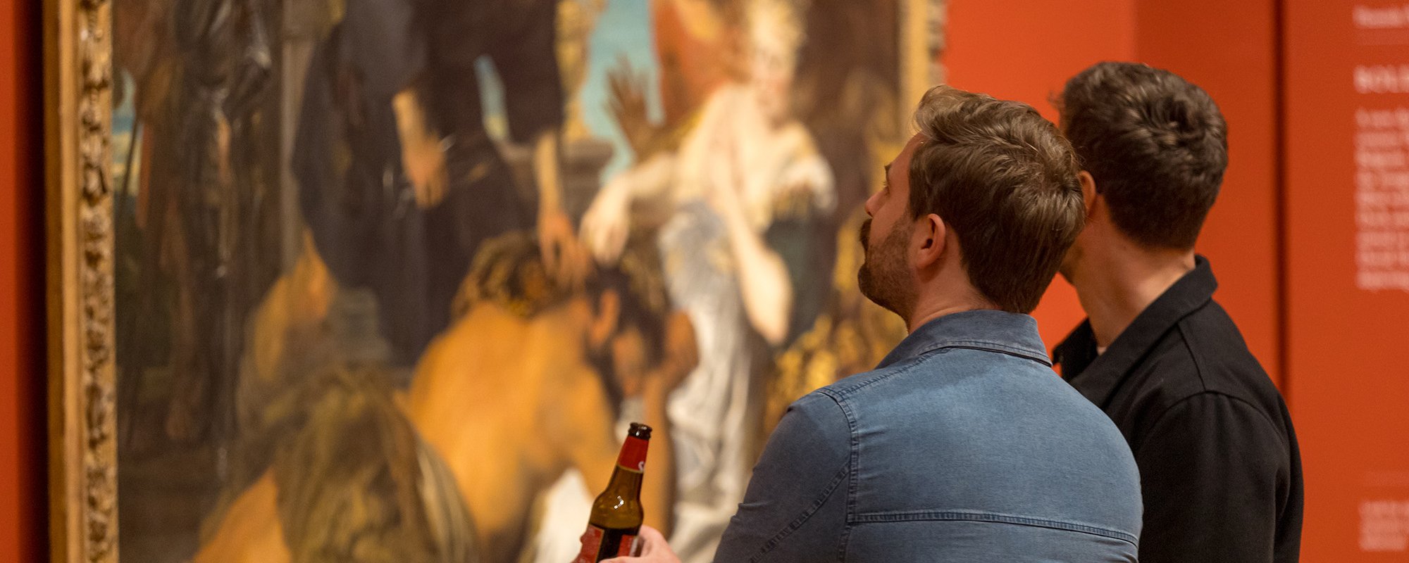Two men closely observing a large classical painting in a museum, one holding a beer bottle in his hand.