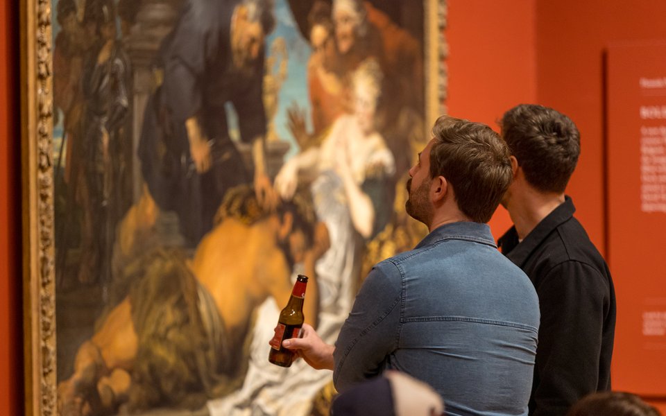 Two men closely observing a large classical painting in a museum, one holding a beer bottle in his hand.
