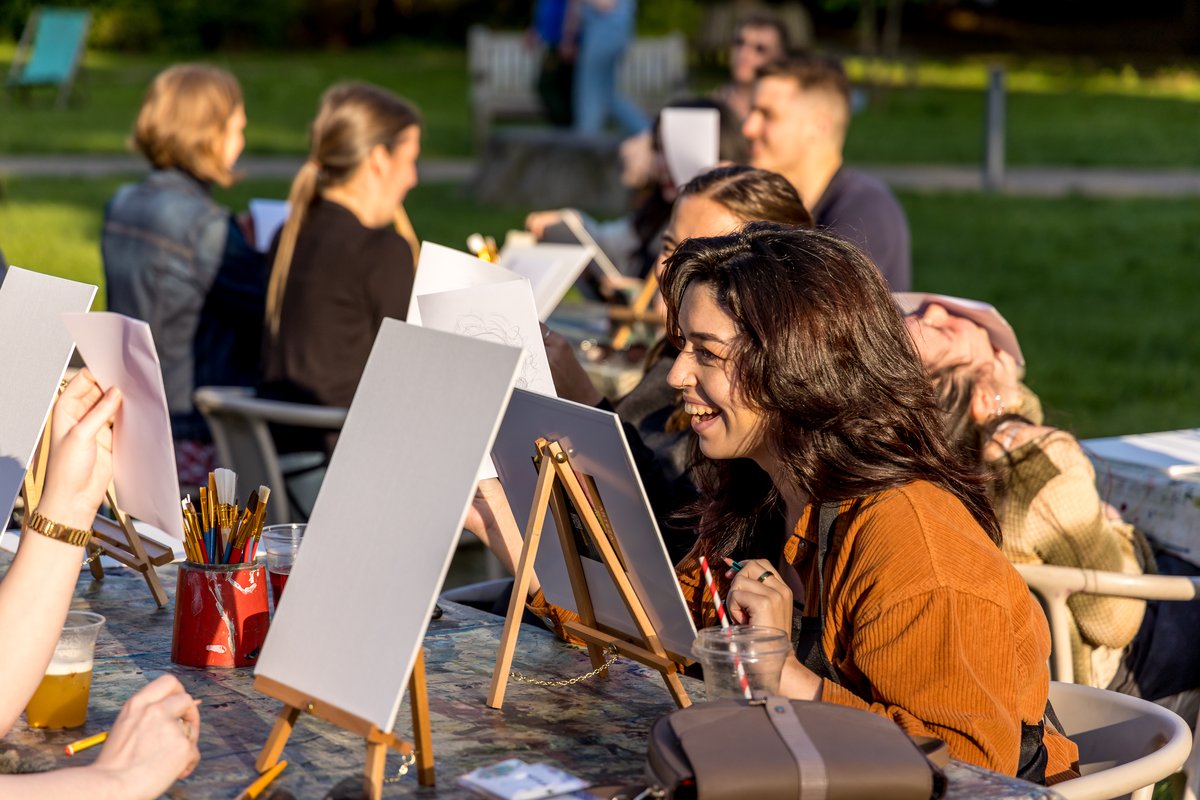 People seated outdoors at a table with easels and blank canvases, preparing to paint in a sunny park setting.