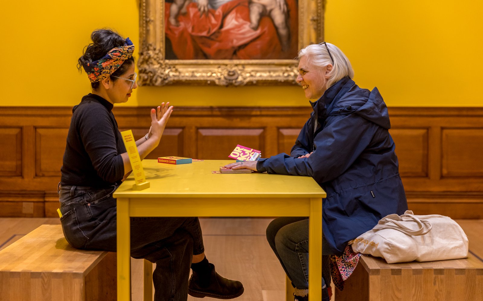 Two women sit at a table talking in a bright yellow room