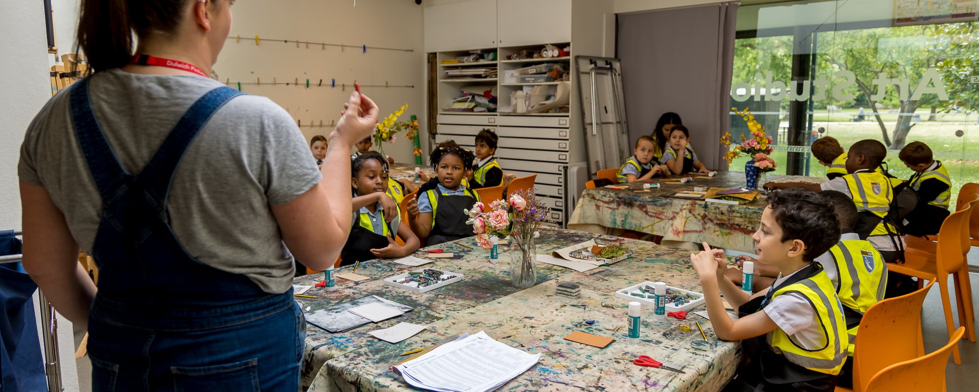A group of young children sit at a table while a woman standing speaks to them
