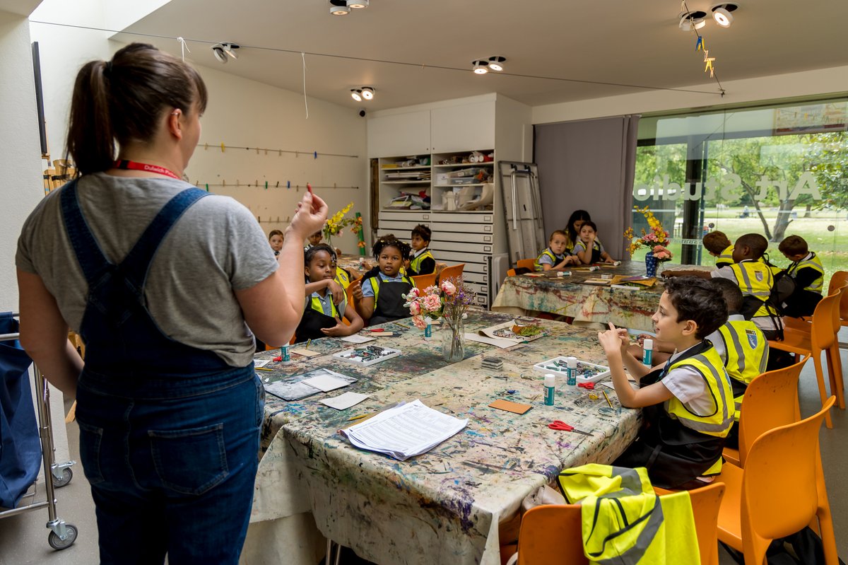 A group of young children sit at a table while a woman standing speaks to them
