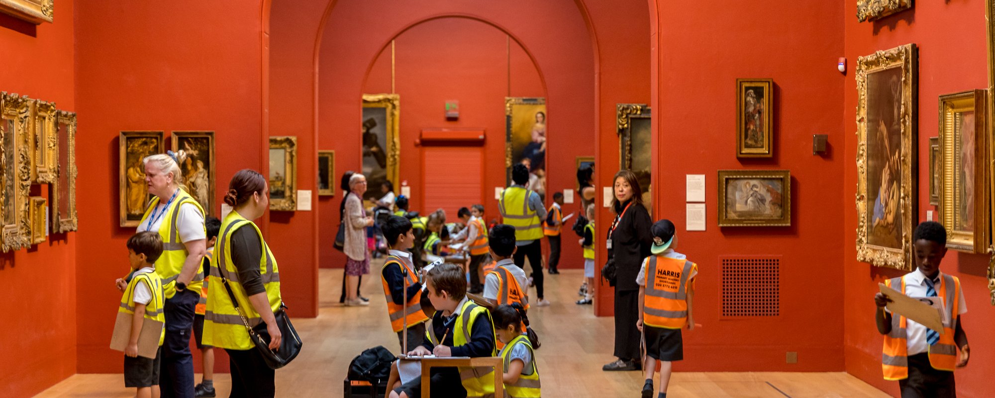 Dulwich Picture Gallery's main red painted gallery space full of children in hi vis yellow and orange vests looking around.