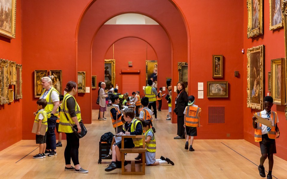 Dulwich Picture Gallery's main red painted gallery space full of children in hi vis yellow and orange vests looking around.