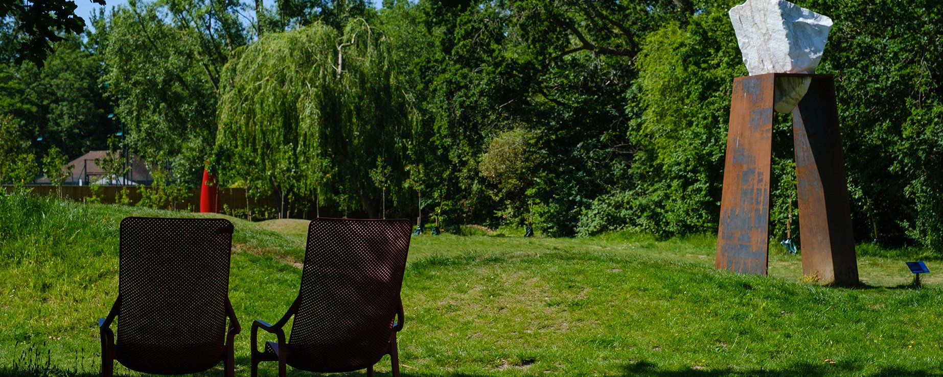 A sculpture with a large white block of stone suspended on a frame with two chairs next to it in a grassy garden