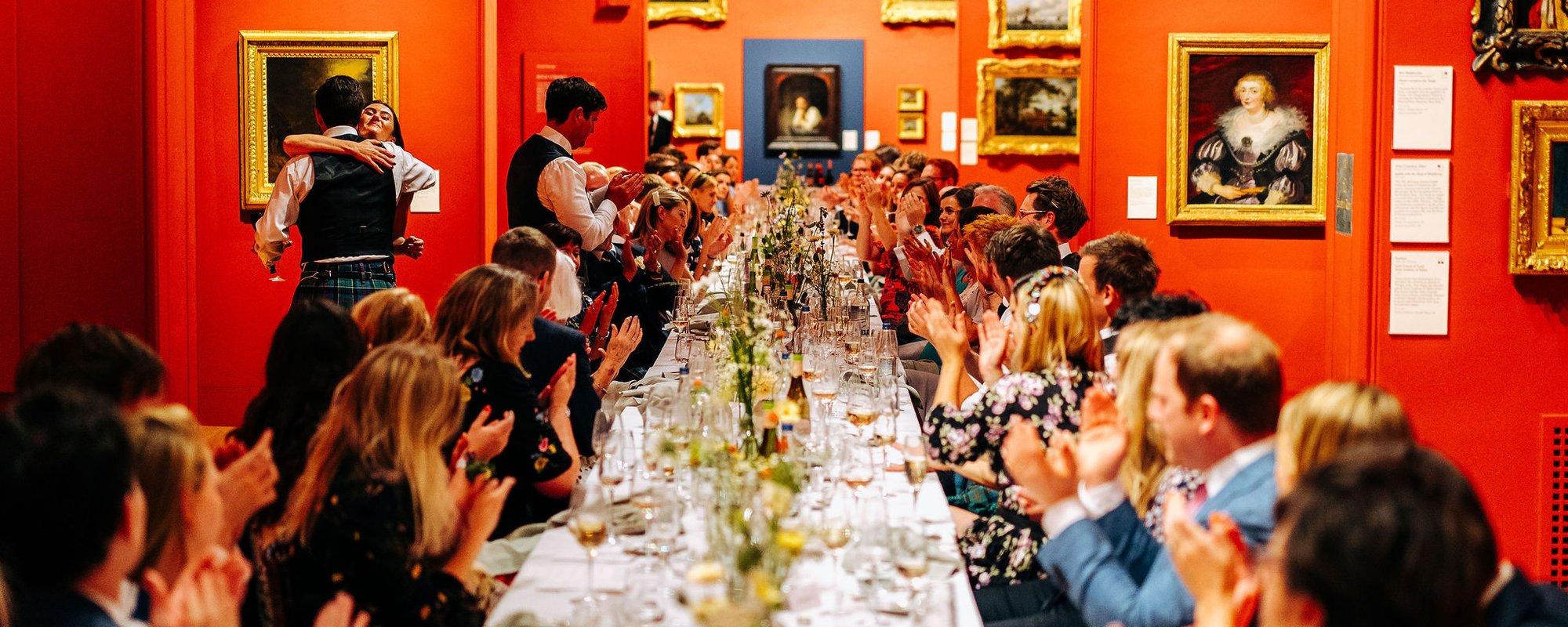 A large banqueting table runs the length of Dulwich Picture Gallery with people either side smartly dressed and clapping. The table is full of floral arrangements and glasses and around them are the walls hung with gold framed paintings.