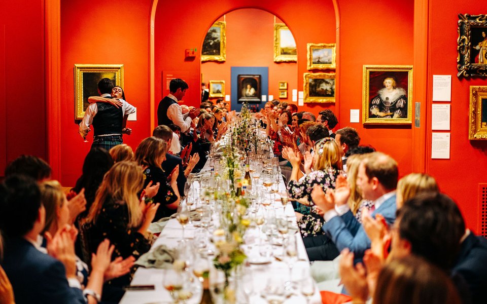 A large banqueting table runs the length of Dulwich Picture Gallery with people either side smartly dressed and clapping. The table is full of floral arrangements and glasses and around them are the walls hung with gold framed paintings.