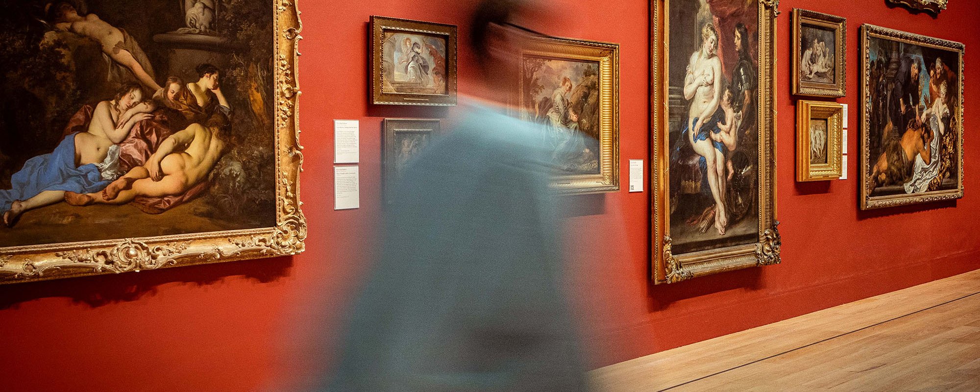 A blurred figure walks right across a gallery space hung with ornate gold framed oil paintings