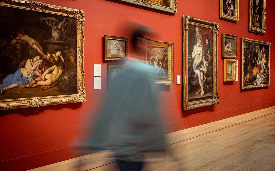 A blurred figure walks right across a gallery space hung with ornate gold framed oil paintings