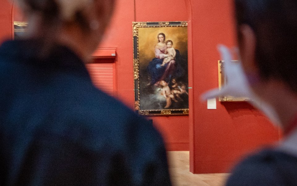 Two blurred figures in foreground look towards a large elaborately framed painting of a Madonna and child.