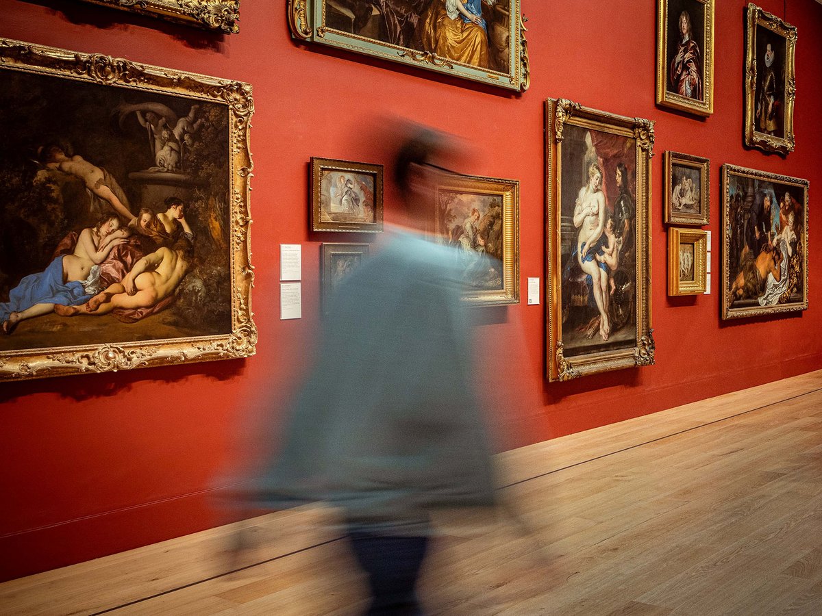 A blurred figure walks right across a gallery space hung with ornate gold framed oil paintings