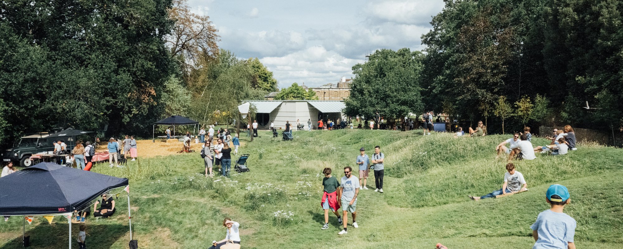 People, including children, are gathered outdoors on a grassy field with trees, tents, and a few structures in the background under a partly cloudy sky.