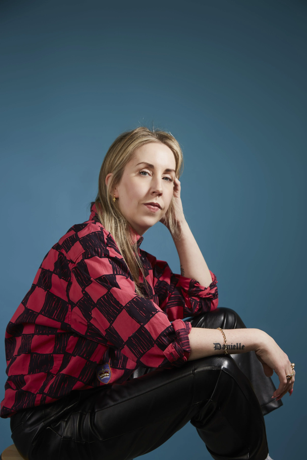Photographic portrait of a woman sat on the floor with back to left of image and legs stretched out right. She wears a black and red cheque shirt with blue jeans.