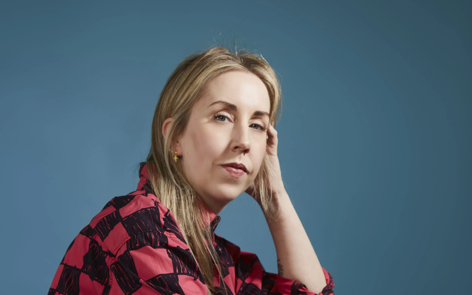 Photographic portrait of a woman sat on the floor with back to left of image and legs stretched out right. She wears a black and red cheque shirt with blue jeans.