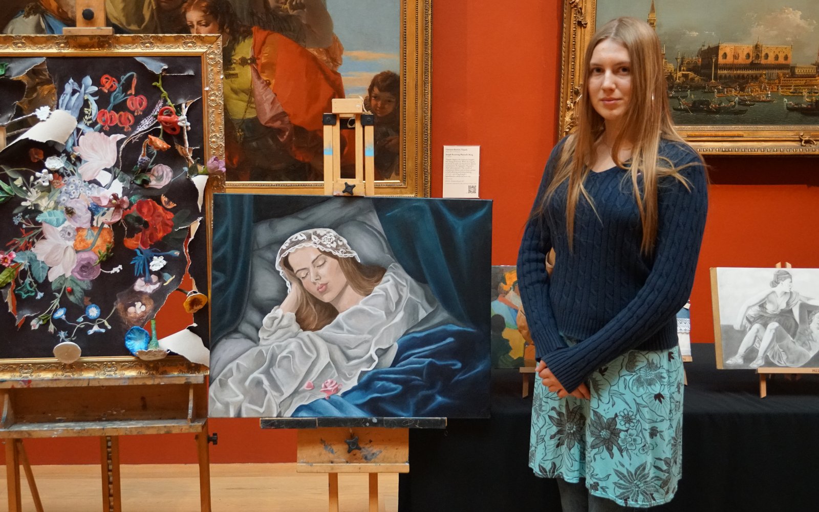 Young girl with long blonde hair stood next to a painting on an easel of a woman sleeping.
