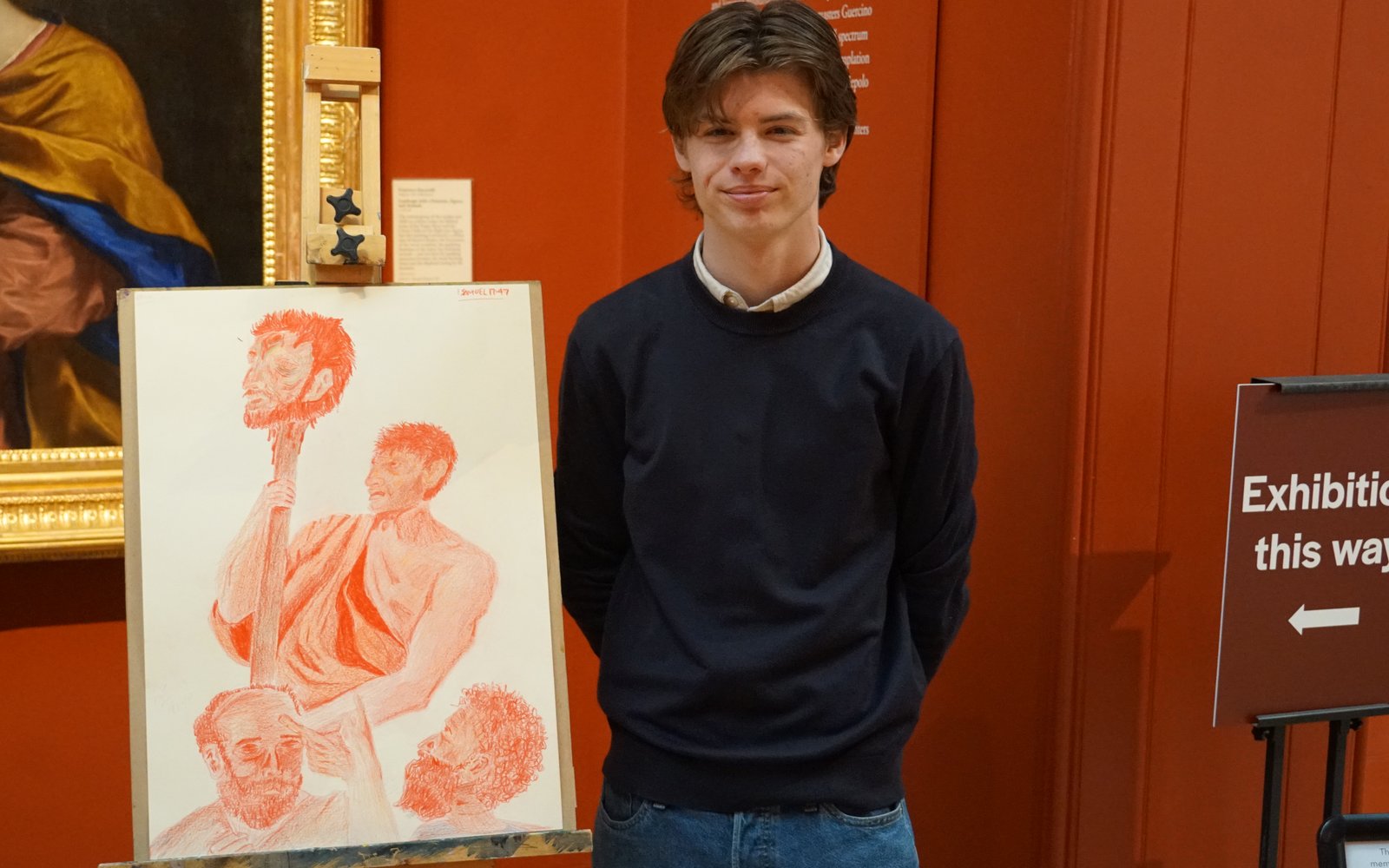 A young man stood next to a sketch on an easel of a man in a toga holding a head on a pike