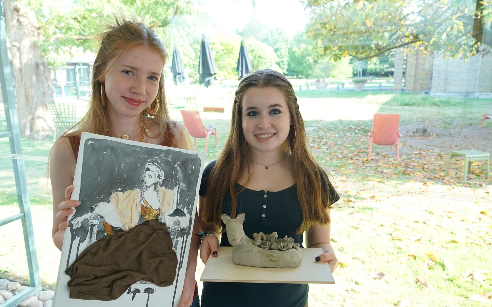 Two young women stood in front of a window one on left holds a collage work and the one right holds a clay sculpture