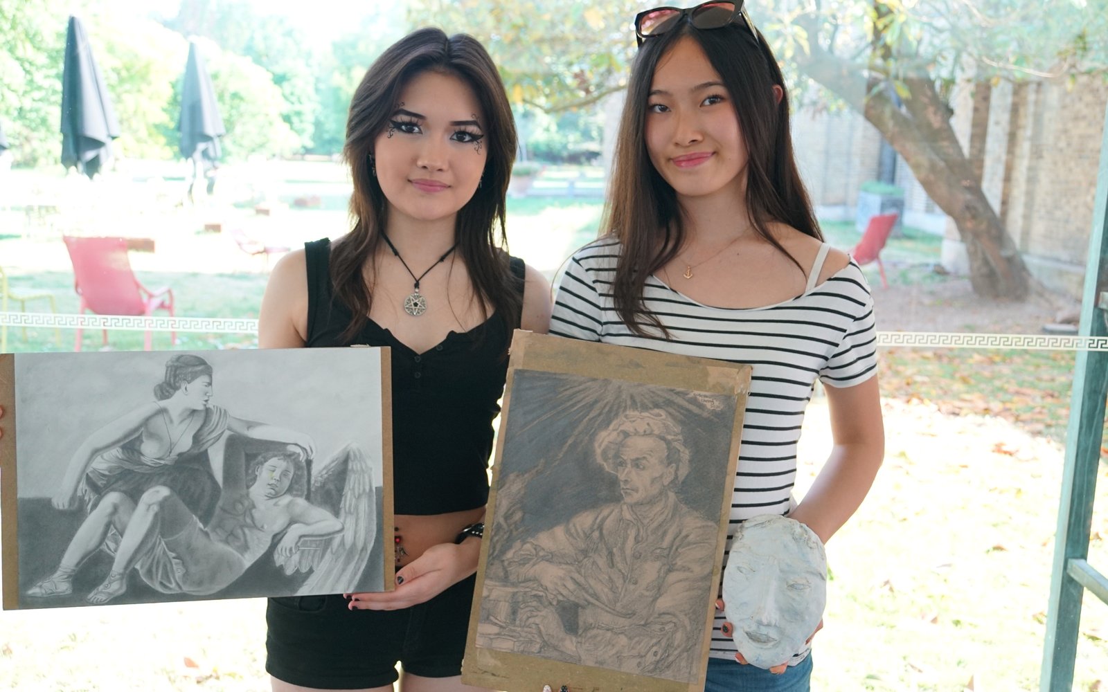 Two young women stood in front of a window both holding skteches.
