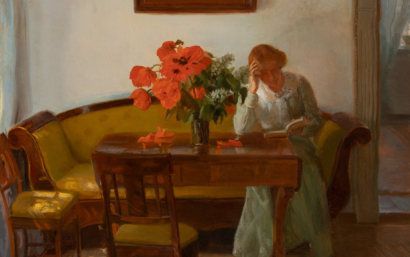 A woman sits at a wooden table with a vase of red poppies on it
