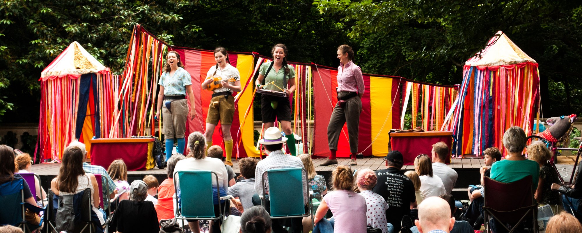A play on an outdoor stage with 4 actors acting with a crowd sat in front.