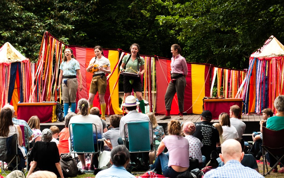 A play on an outdoor stage with 4 actors acting with a crowd sat in front.