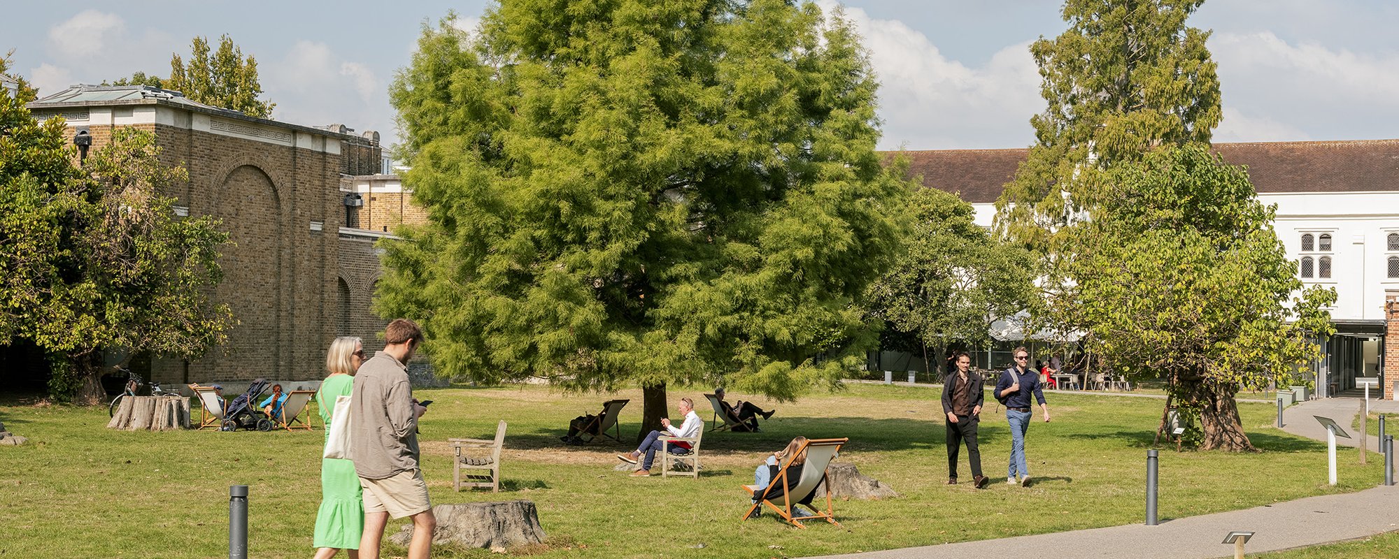 The gardens of Dulwich Picture Gallery with people walking along the paths and across the grass while others sit in chairs and deckchairs on the grass. There are tall leafy trees and you can see the Gallery's brick building in the back.
