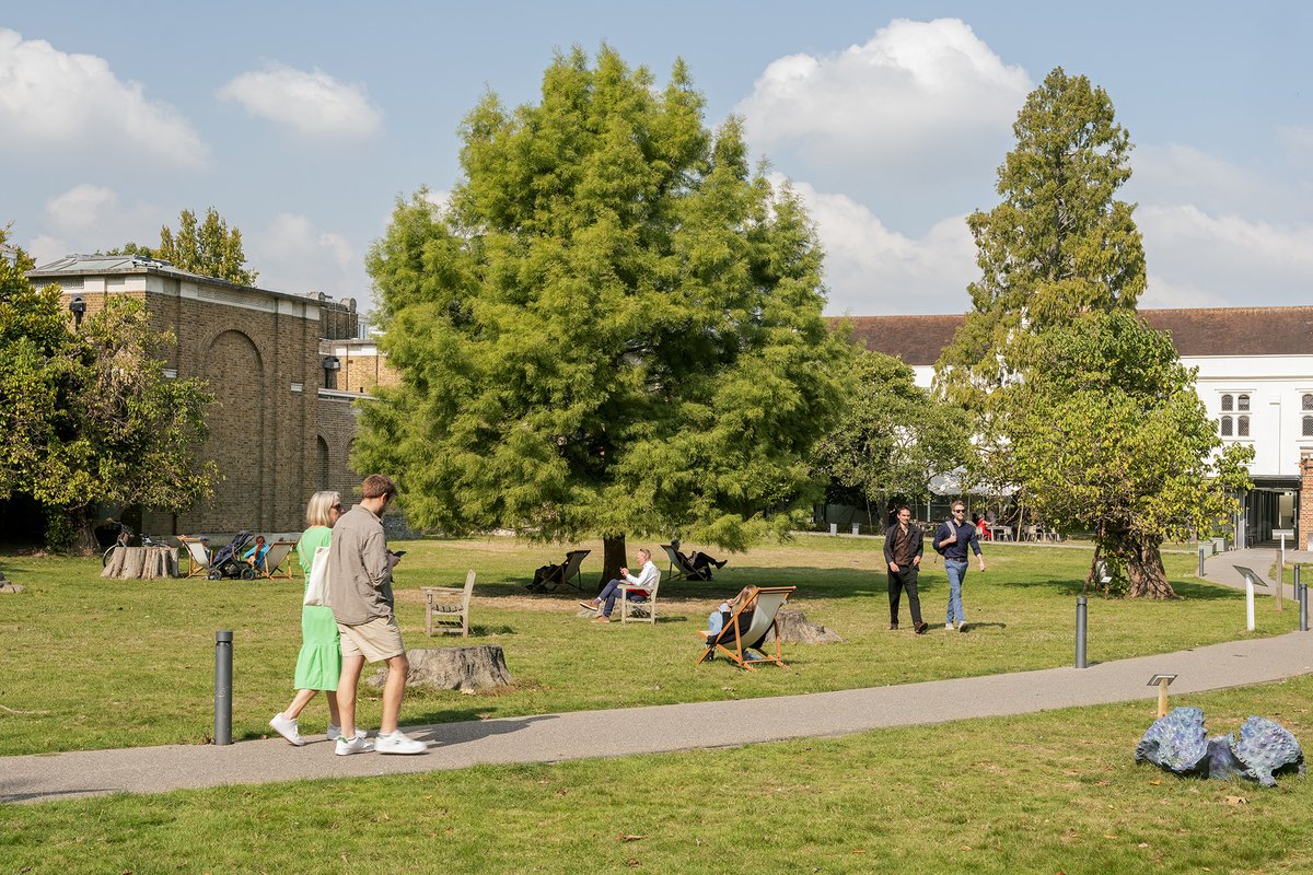The gardens of Dulwich Picture Gallery with people walking along the paths and across the grass while others sit in chairs and deckchairs on the grass. There are tall leafy trees and you can see the Gallery's brick building in the back.