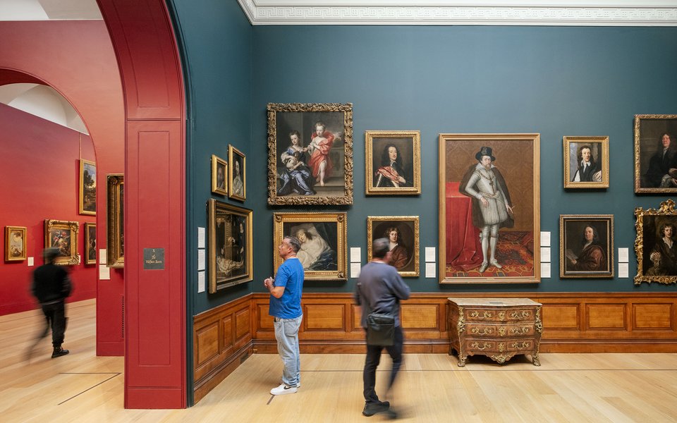 Two men stand looking at portraits in the British Portrait Room at Dulwich Picture Gallery with gold framed portraits hung on a dark blue wall.
