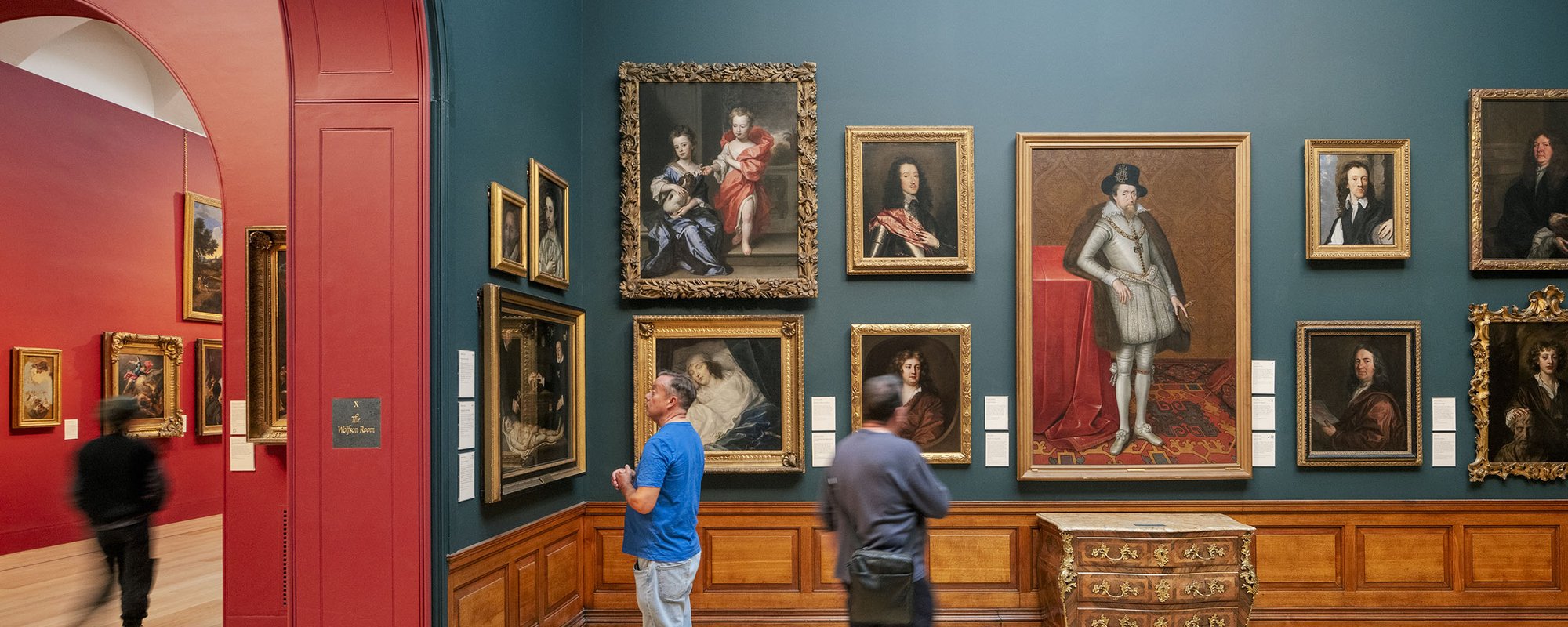 Two men stand looking at portraits in the British Portrait Room at Dulwich Picture Gallery with gold framed portraits hung on a dark blue wall.