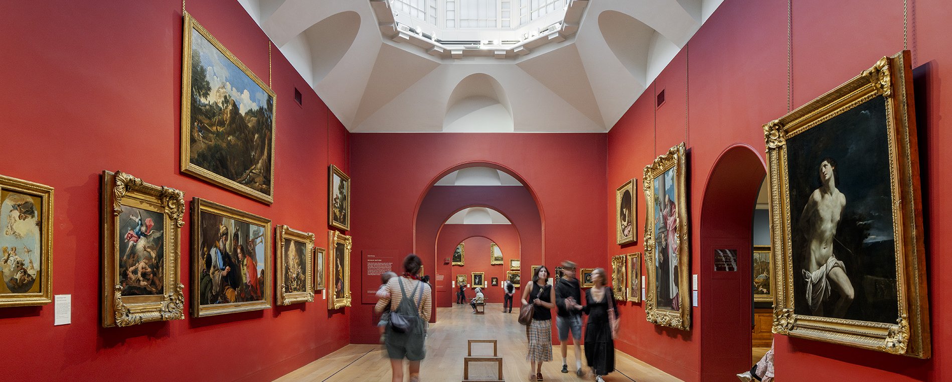 Visitors explore a bright art gallery with red walls adorned by classical paintings in ornate gold frames under a large skylight ceiling.
