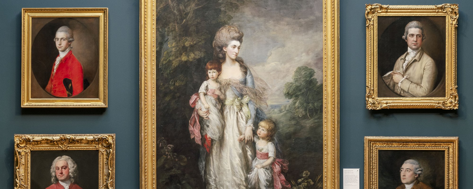 A gallery space with blue walls and a wood paneled bottom section hung with gold framed historic portraits.