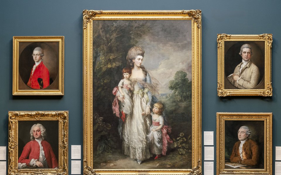 A gallery space with blue walls and a wood paneled bottom section hung with gold framed historic portraits.